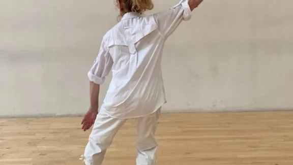 A photograph from behind of Leslie Satin dancing in a white collared shirt and white pants.